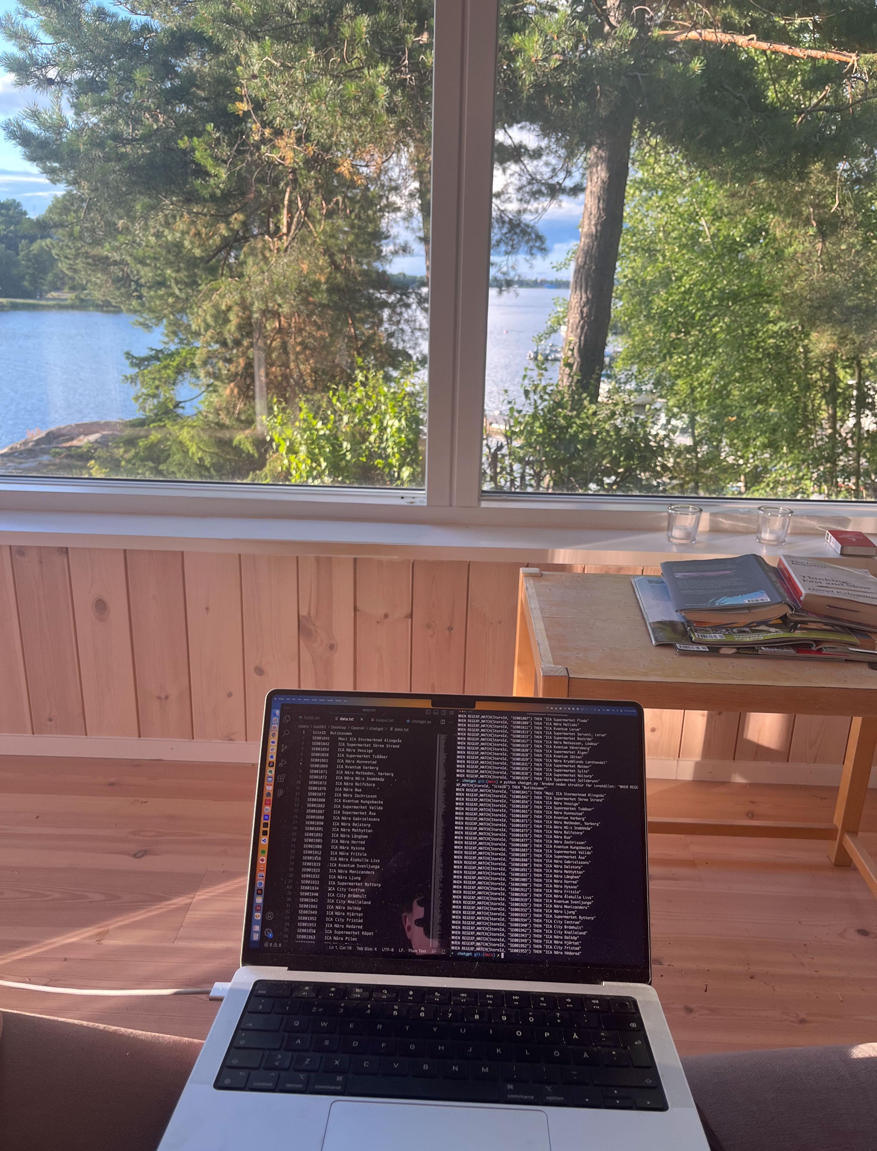 A laptop open on a table in front of a window with a view of a lake and trees.