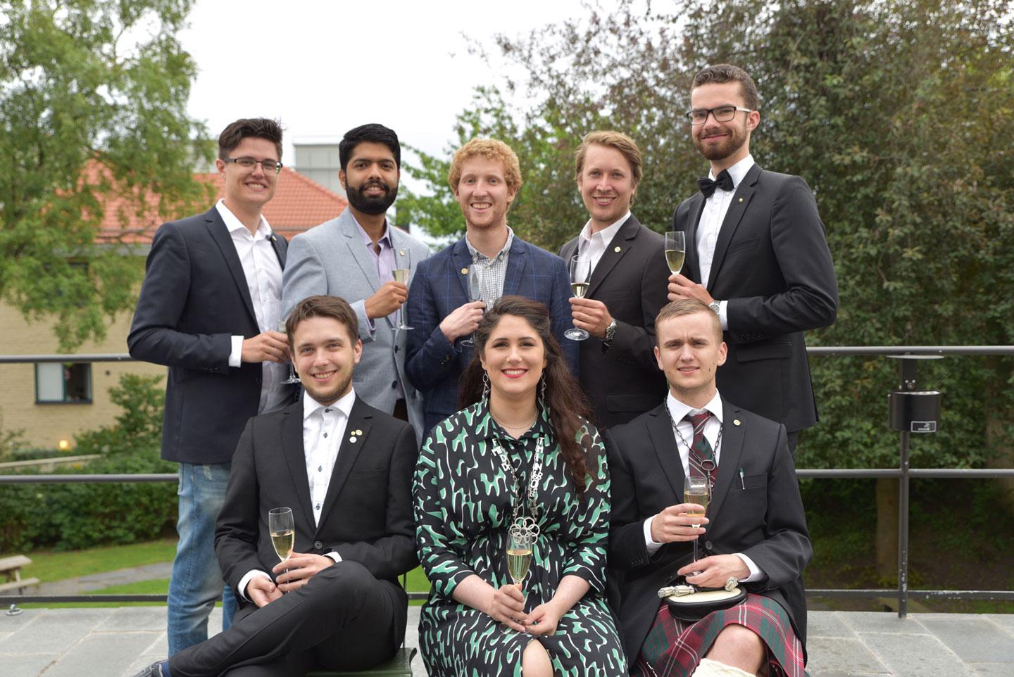 Group of smiling people dressed in party attire posing outdoors with champagne glasses.