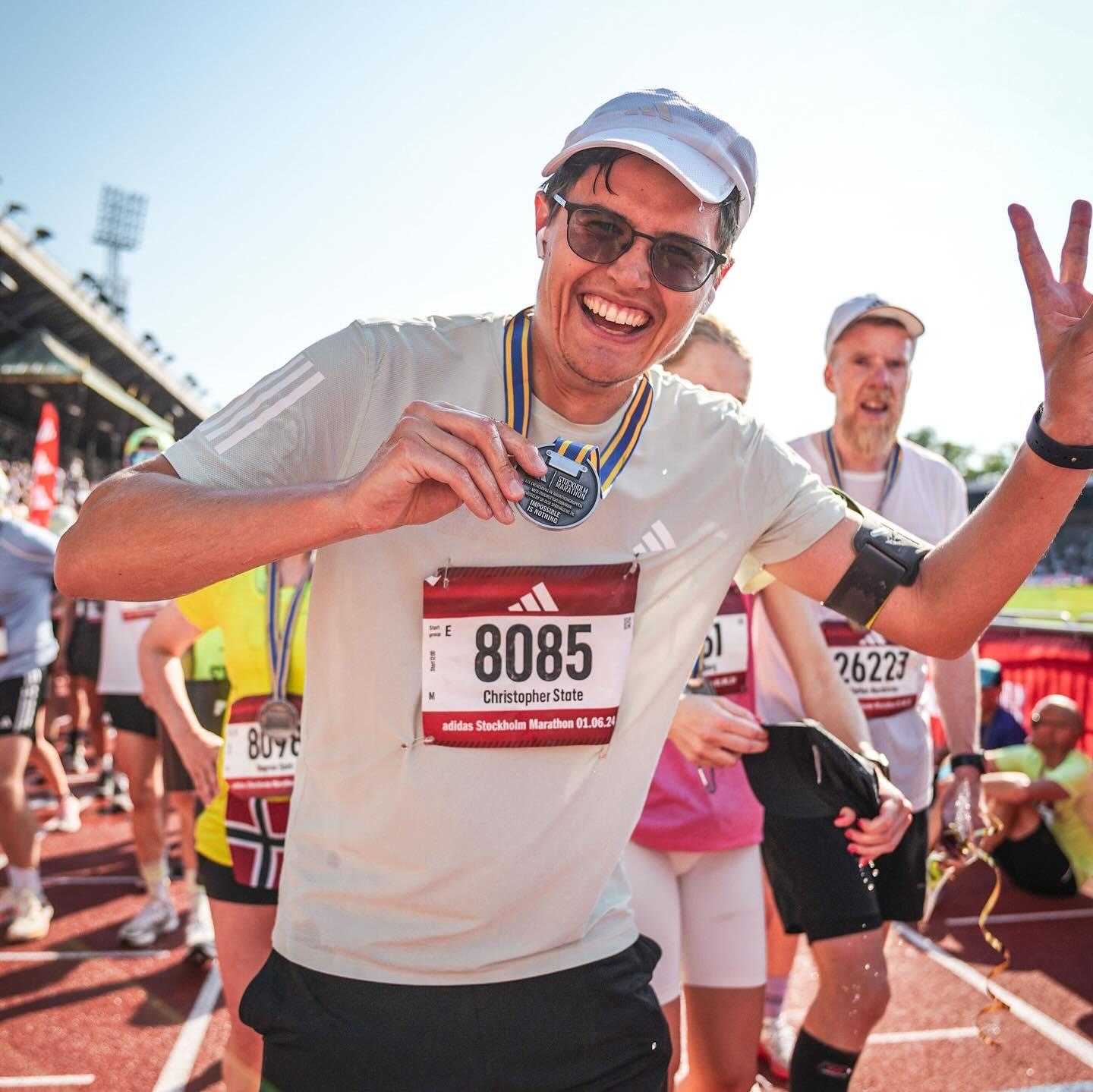 A smiling marathon runner, Christopher State, holds his medal proudly at the finish line of the Adidas Stockholm Marathon. He is wearing sunglasses, a white cap, and a light-colored shirt with the race number 8085. Other runners and spectators are visible in the background under a clear sky.