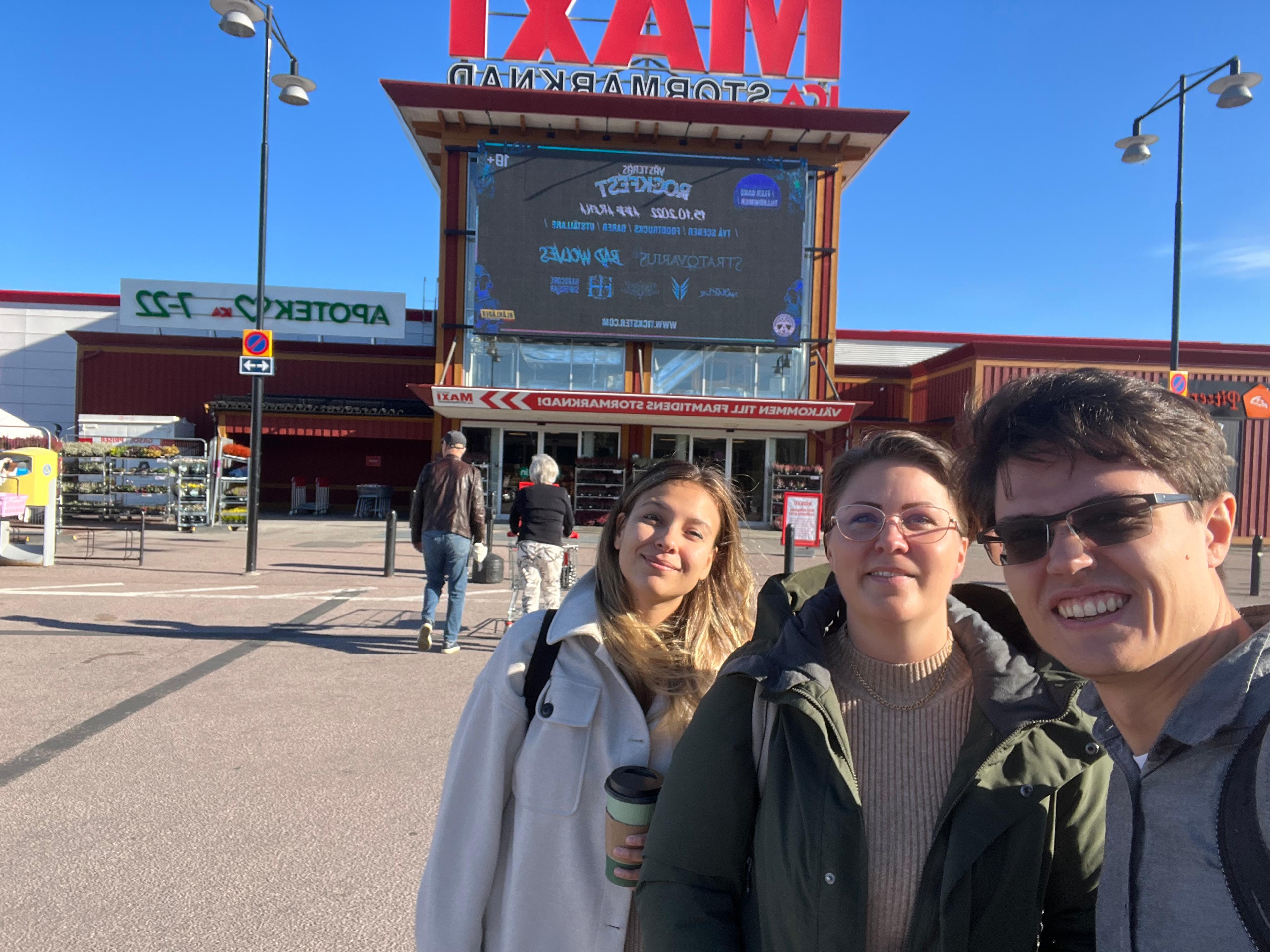 Three people are smiling at the camera in front of a cinema entrance on a sunny day.