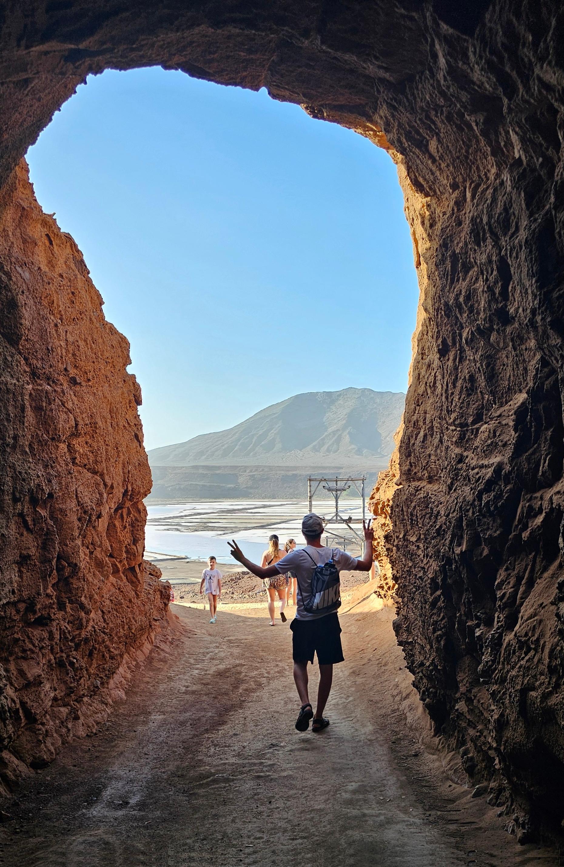 A person standing in a cave entrance with arms outstretched towards a beach and mountains in the background.