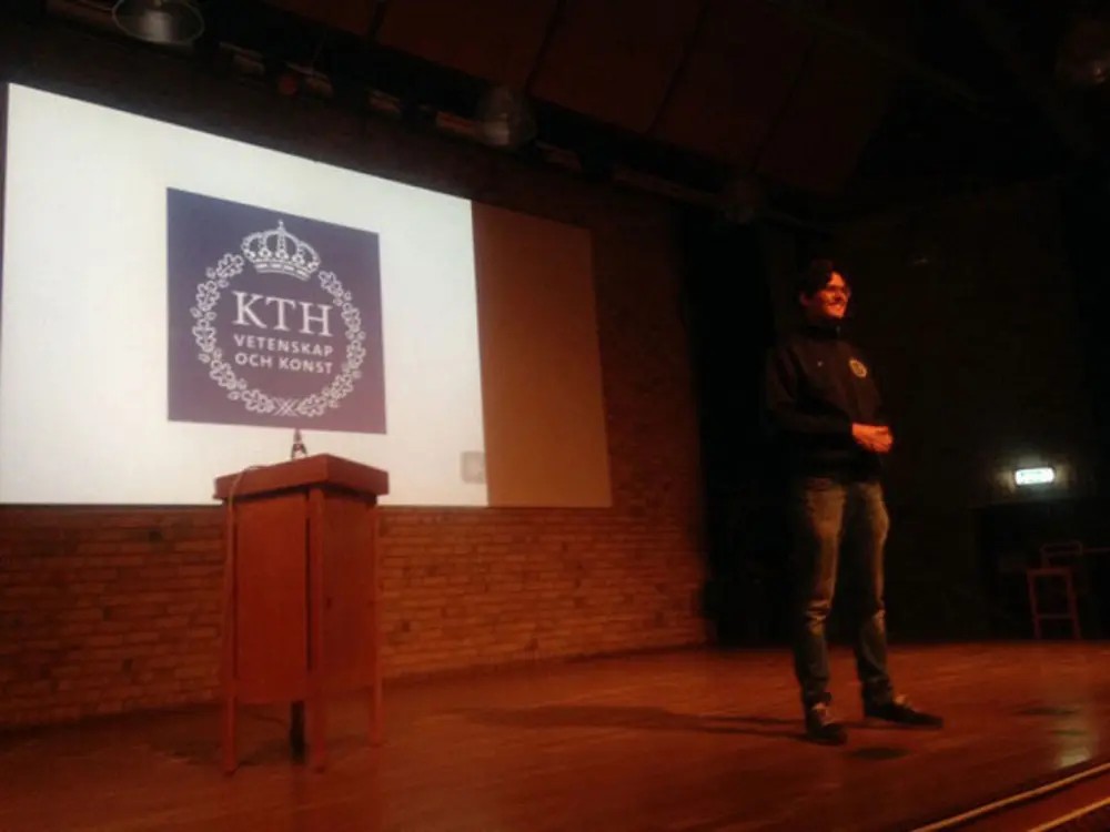 A person stands at a lectern in a dark lecture hall with a projected KTH logo on a screen in the background.