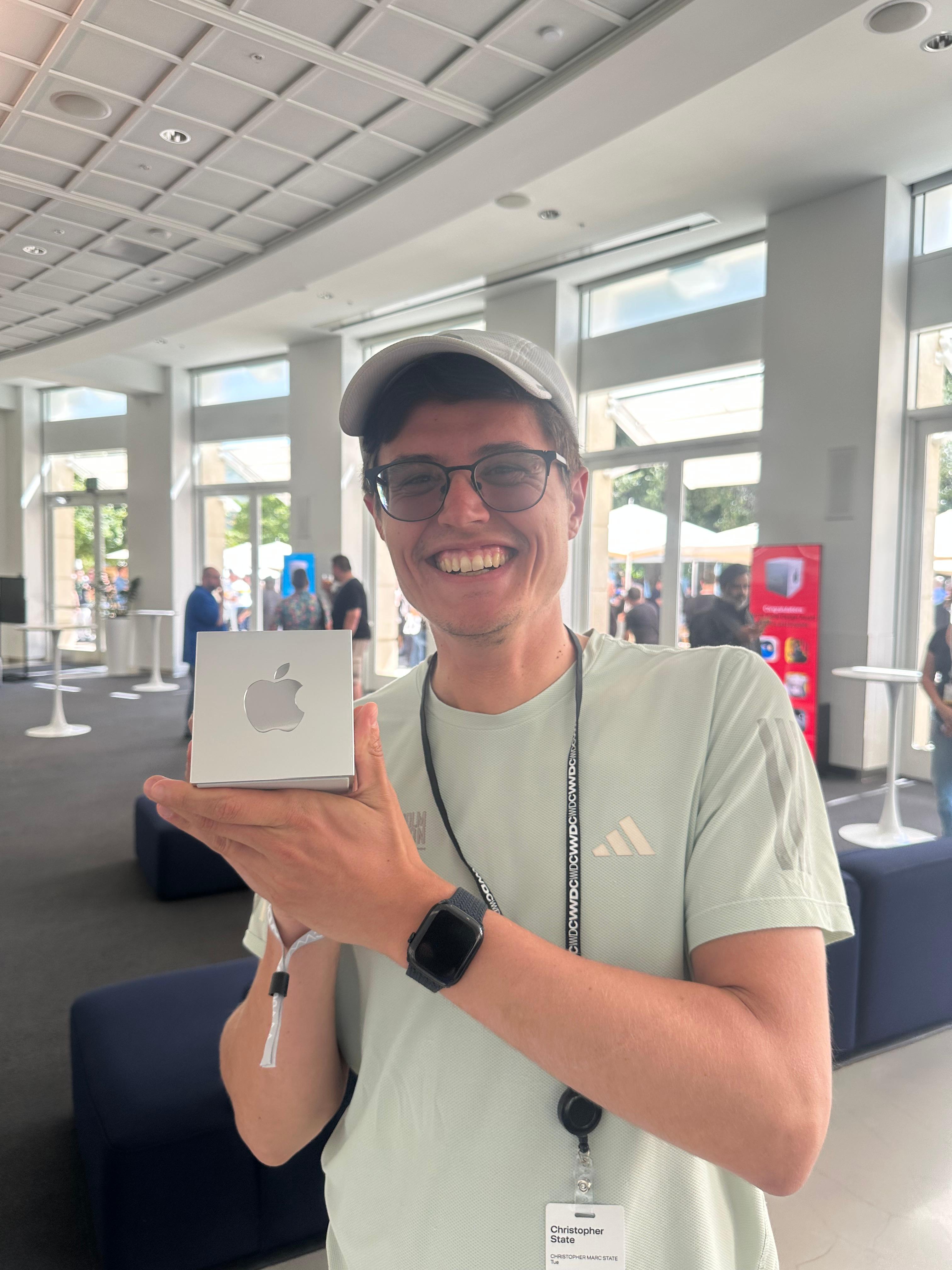 A young man smiles while holding a small Apple product box in a modern indoor space with large windows. He is wearing a light green shirt and a cap.