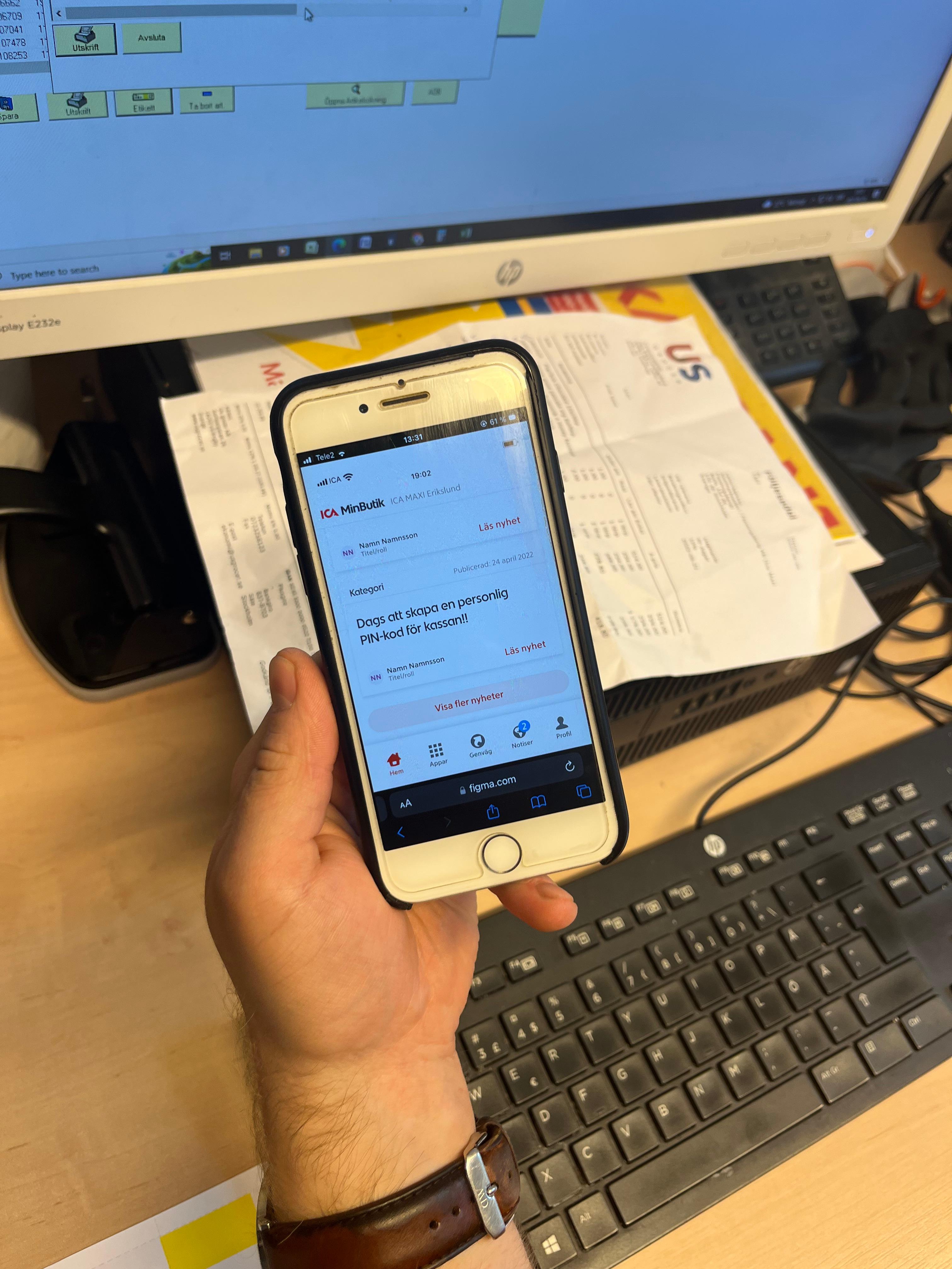 A person holds a smartphone with an open intranet app in front of a computer screen and a keyboard on a desk.