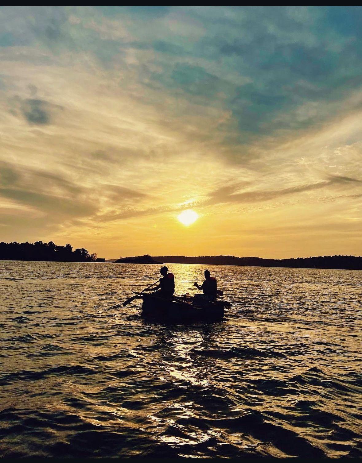 Two people sit in a small raft on a lake at sunset, with vibrant colors in the sky reflecting on the water. The scene captures a peaceful moment in nature.