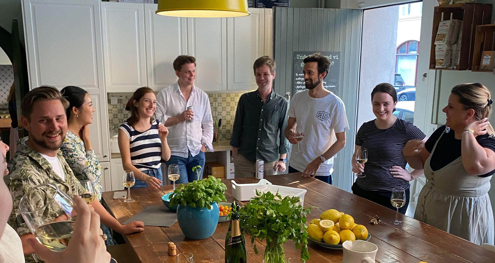 A group of people gathered in a kitchen with food and drinks on the table, appearing to be in a social gathering.